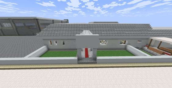 Foco em uma segunda entrada da escola. Esta entrada possui uma cobertura de madeira sustentada por quatro pilares finos. Debaixo da cobertura, há dois bancos de madeira voltados um para o outro, caminho de tapete vermelho entrada dos alunos.