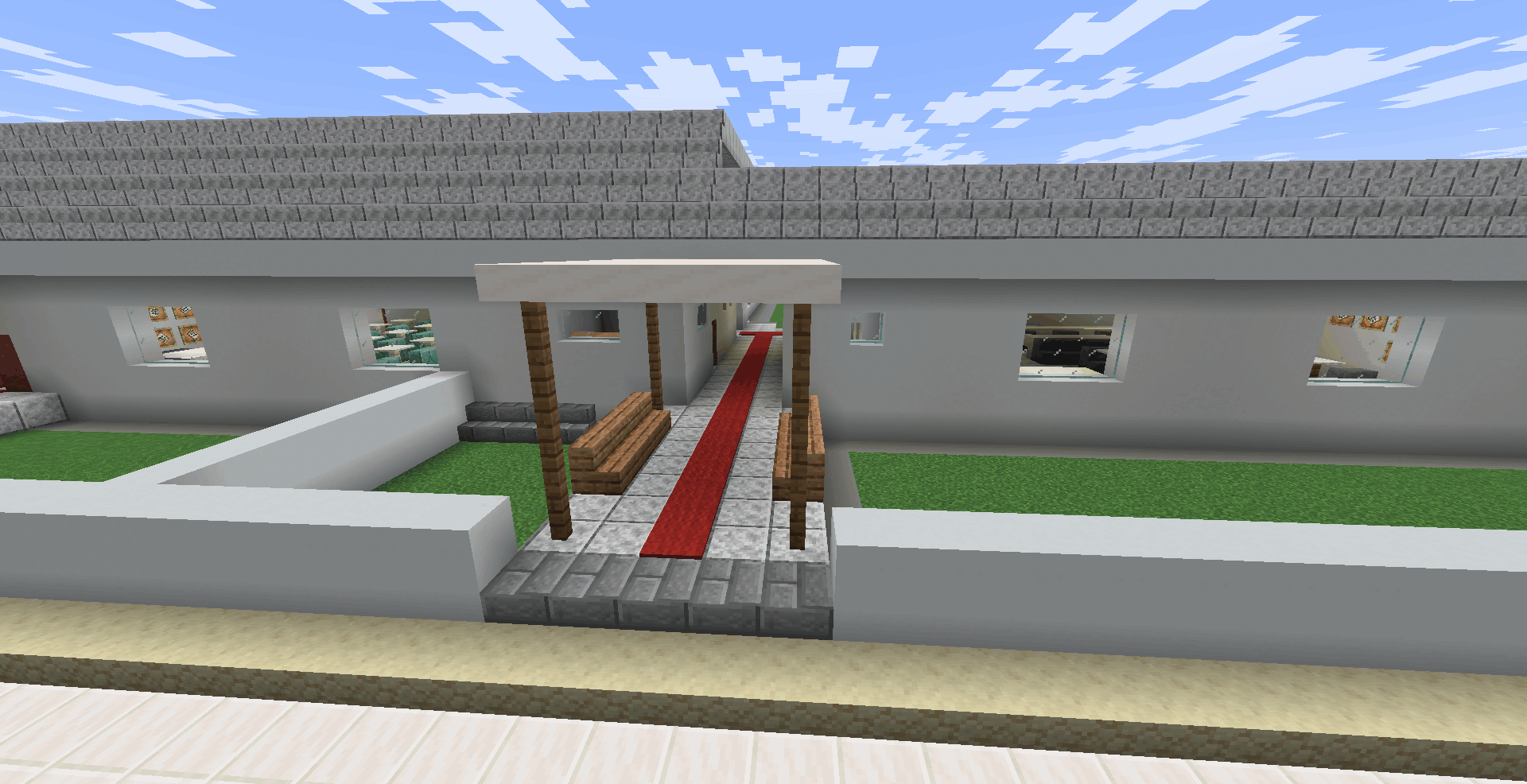 A imagem mostra a frente da escola. É um prédio térreo longo, construído com blocos brancos e telhado cinza. No centro, há uma entrada com um pequeno telhado suspenso. Um caminho de blocos cinzas com um tapete vermelho centralizado leva até a porta. À frente da escola, há dois gramados verdes retangulares cercados por muretas brancas baixas.