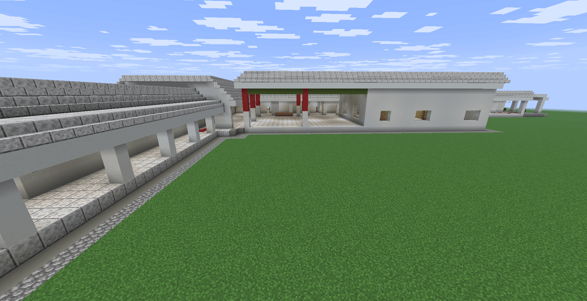 Vista de uma área aberta dentro da escola (pátio). À esquerda, há um corredor coberto com colunas brancas e chão de pedra clara. Ao fundo, uma área de convivência ampla com pilares detalhados em vermelho e verde. O chão é de um piso quadriculado cinza claro.