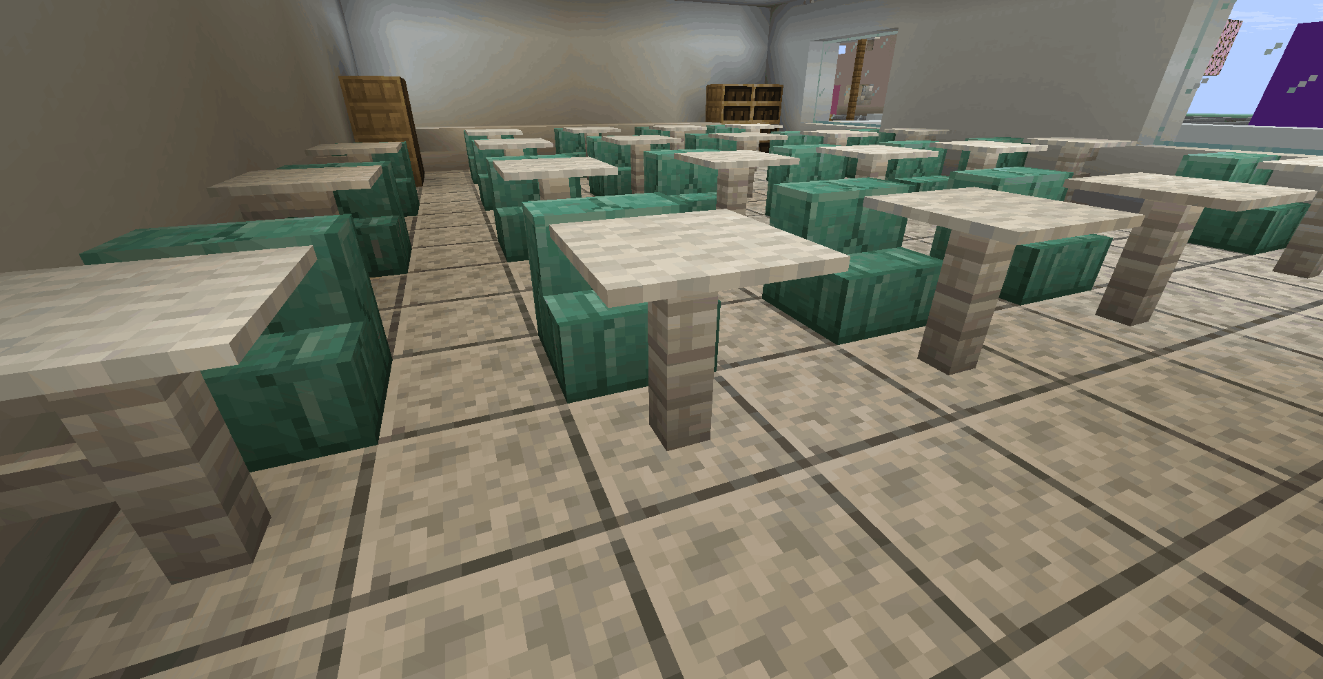 Vista de dentro de uma sala de aula. O ambiente é organizado com várias fileiras de mesas brancas individuais. Cada mesa tem uma cadeira de blocos verdes (prismarinho). Ao fundo, vê-se uma estante de livros de madeira e uma porta fechada.