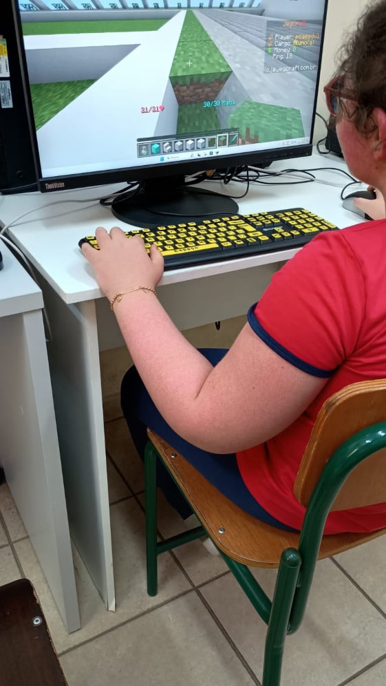 Uma estudante está sentada em uma mesa de computador jogando Minecraft. Ela usa uma camiseta vermelha e utiliza um teclado especial com teclas amarelas grandes e letras pretas, projetado para facilitar a visualização. Na tela do monitor, vê-se o ambiente do jogo em construção, com blocos de grama e áreas cinzas.